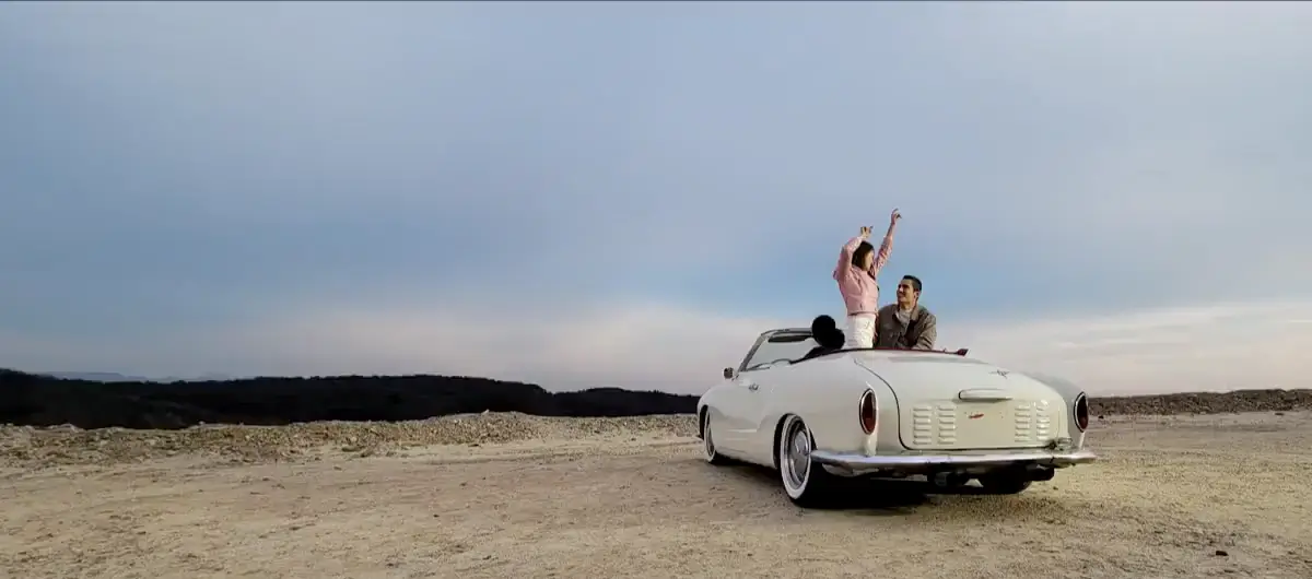At 1x: a white convertible on a desert track, friends standing up in the back