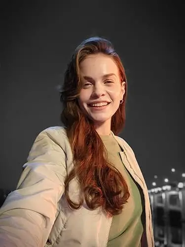 A smiling woman with long red hair takes a selfie at night, city lights blurred behind her