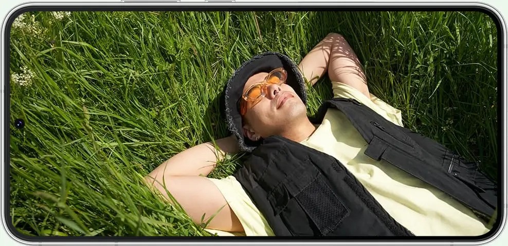 Galaxy S22+ held sideways, showing a man in sunglasses lying back in long grass
