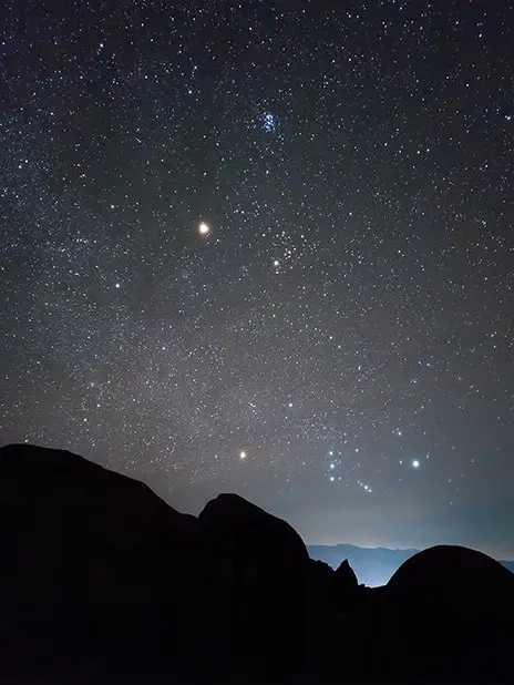 A starry night sky over the dark silhouette of rocky hills
