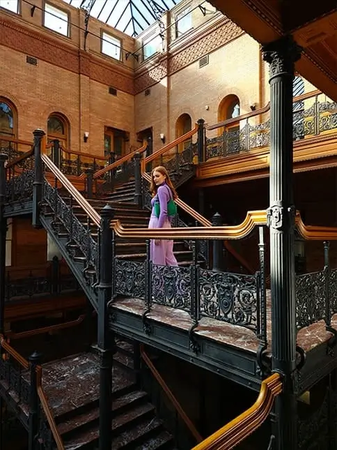 A woman on a landing of a brick atrium with tiers of ornate iron staircases, lit by a glass roof