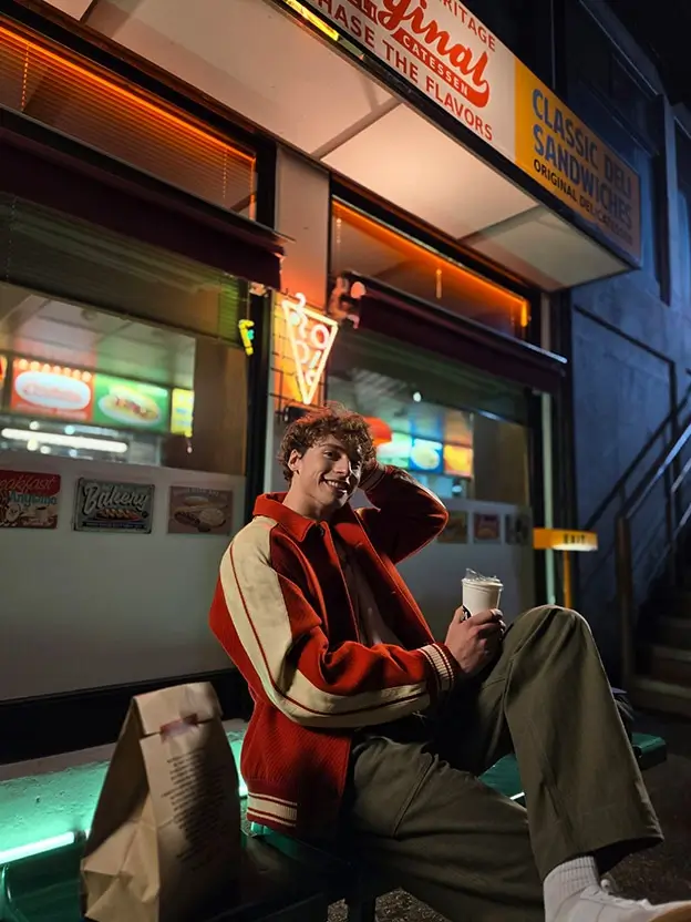 A young man in a red jacket sitting outside a neon-lit takeaway at night