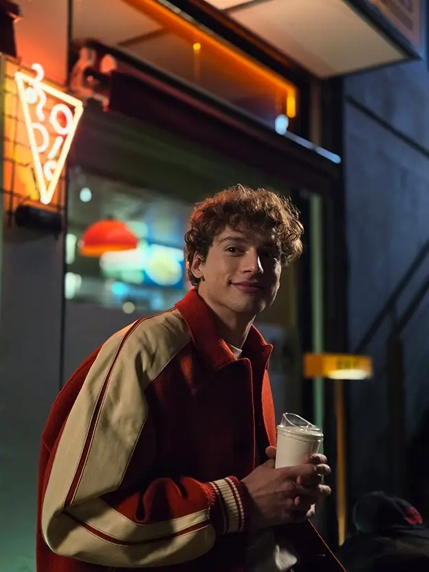 Closer: the young man smiling, holding a cup, a neon pizza sign behind him