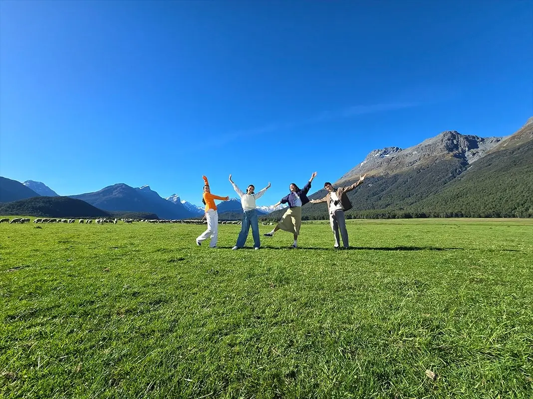 At 0.6x: a wide green field under blue sky and mountains, four friends small in the middle