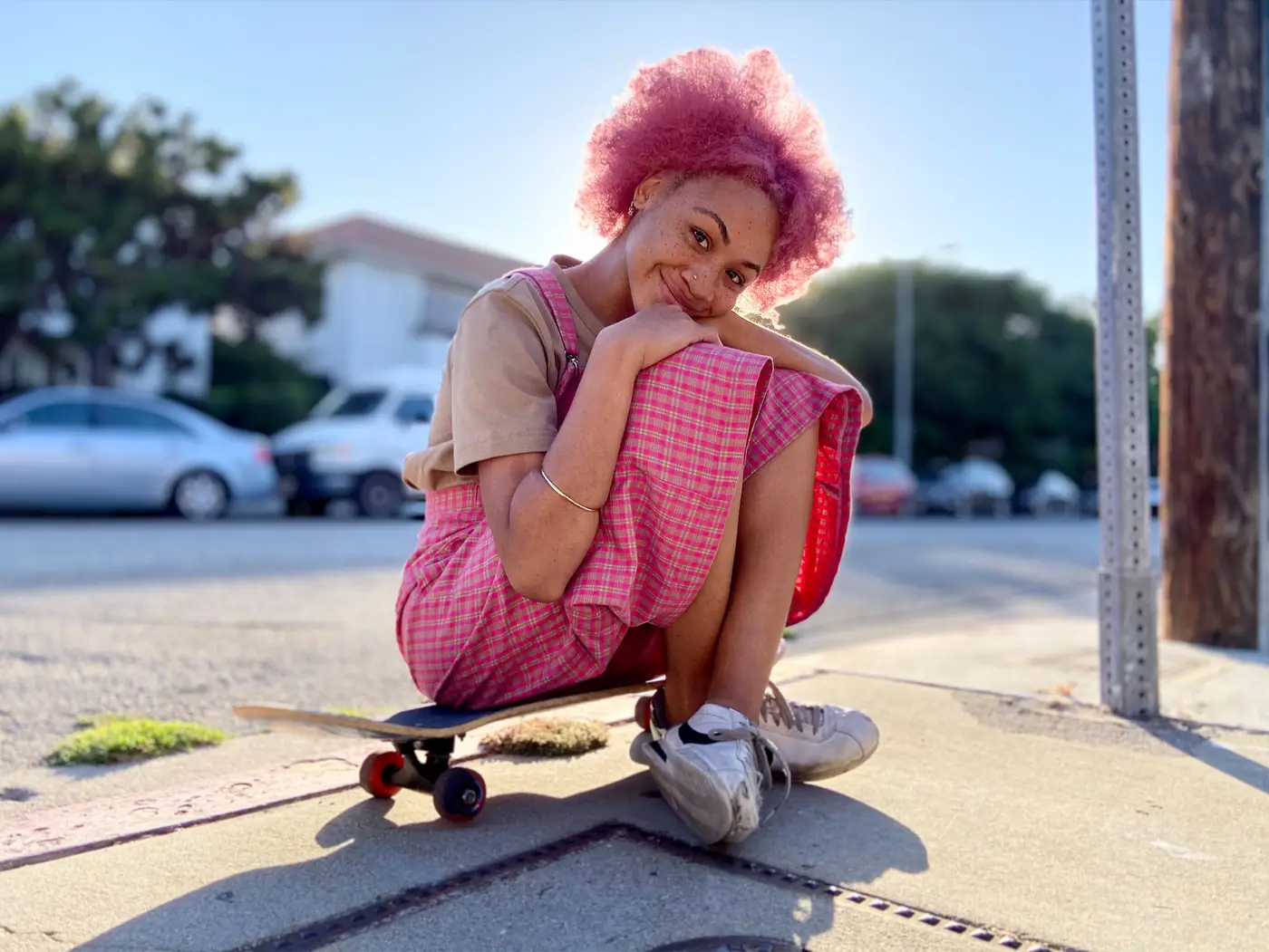 A person with pink hair sitting on a skateboard on a sunny street