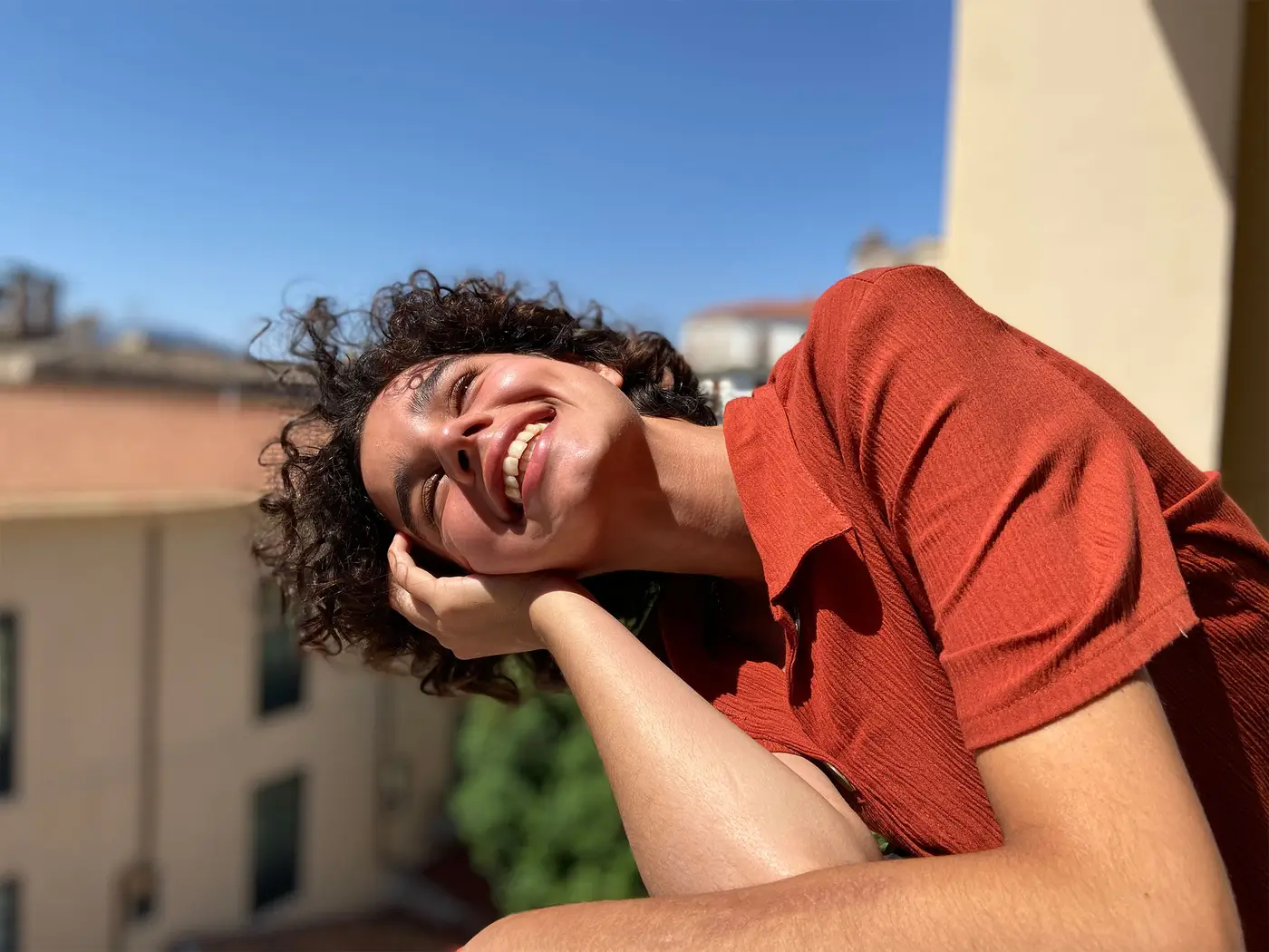 A smiling person in a red shirt resting their head on their hand in bright sun