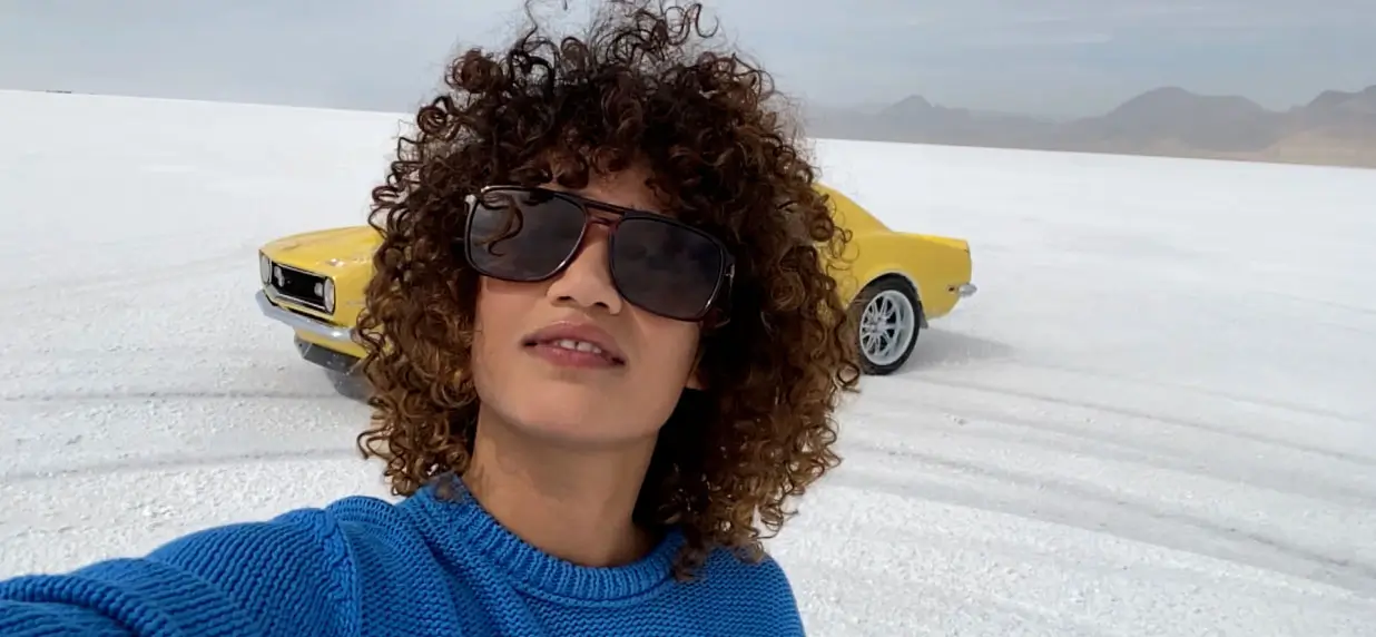 A wide selfie of a person in sunglasses and a blue jumper, with a yellow classic car behind on a salt flat