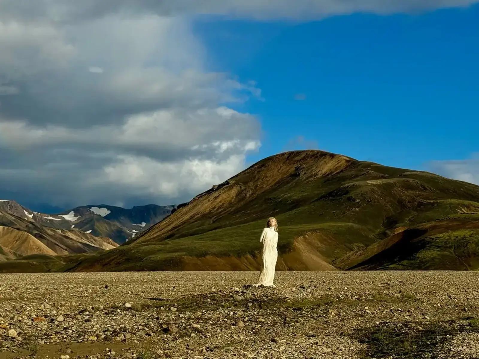 At 65 mm the person in a long white dress stands against a green hill under the clouds