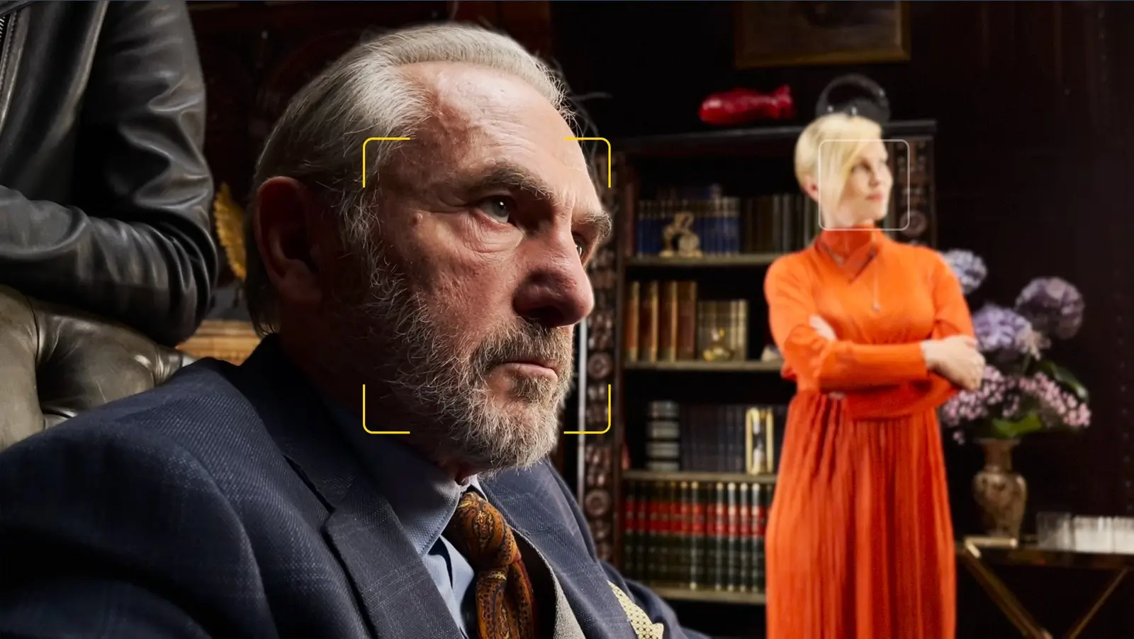 An older person with a grey beard in sharp focus in a dark study, with a person in an orange dress blurred by the bookshelves behind