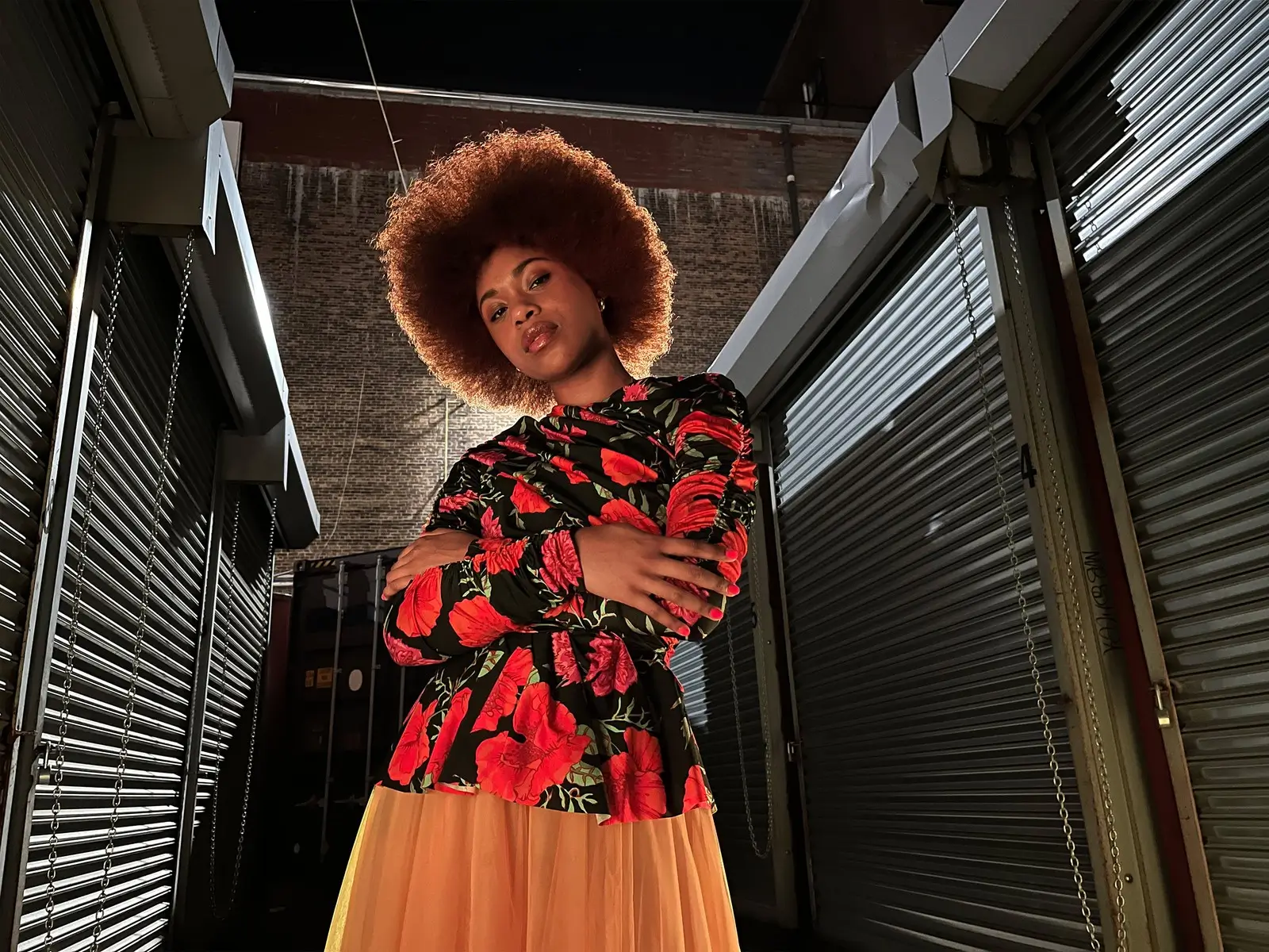 A person in a red and black outfit posing between metal shutters on a city street at night