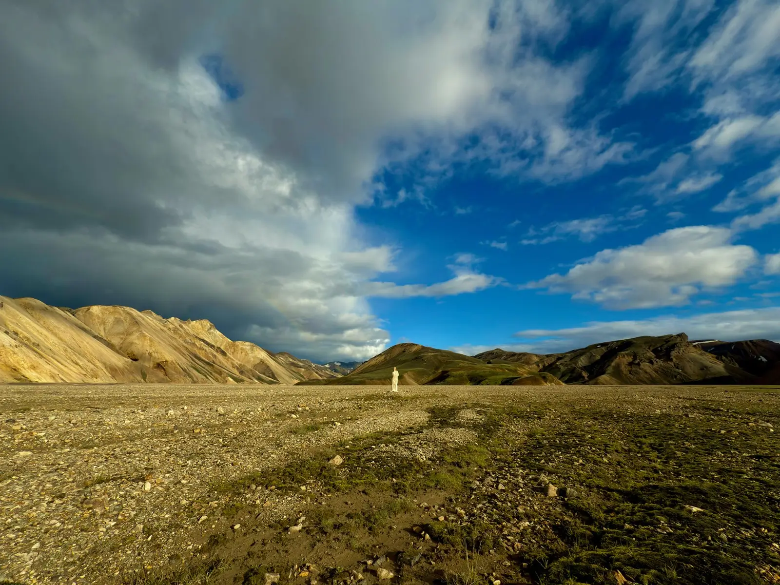 A stony plain running to striped mountains under heavy cloud, a person in white tiny at the centre, at 0.5x