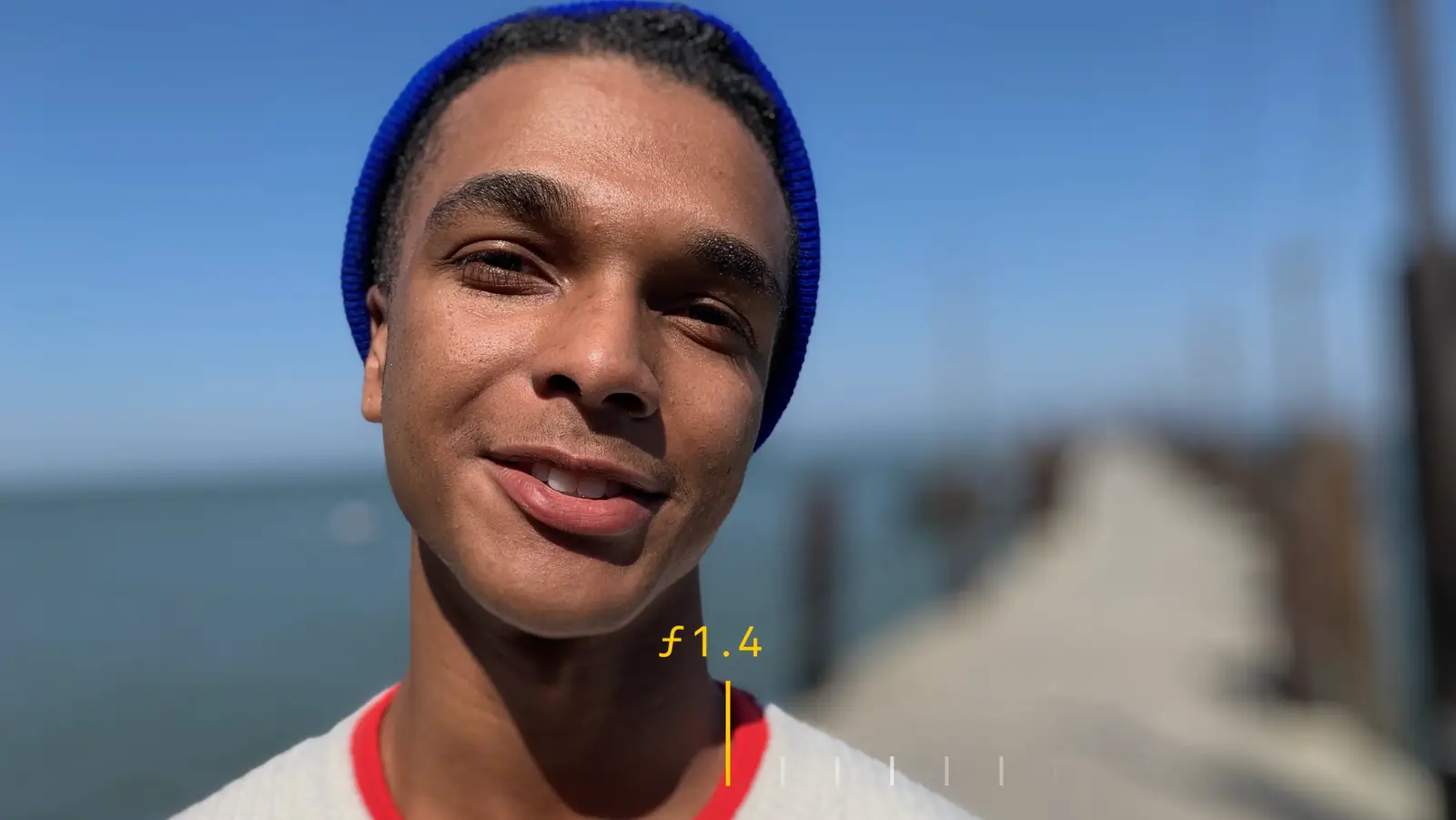 A smiling man in a blue beanie on a pier, the pier and sea heavily blurred behind him