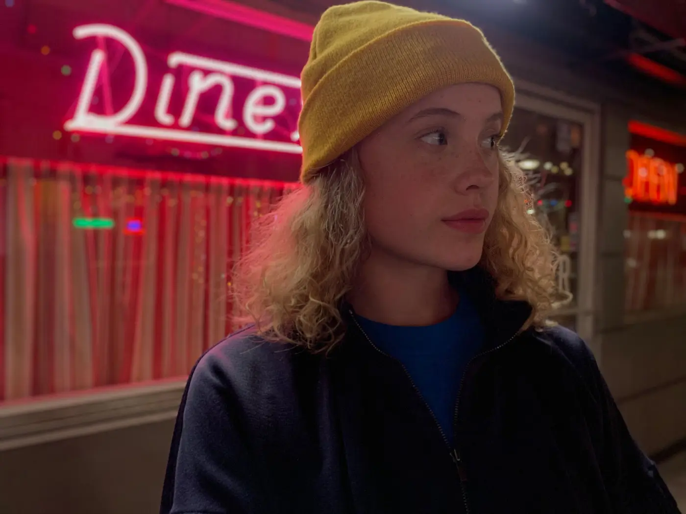A girl in a yellow beanie outside a diner at night, its pink neon sign blurred behind her