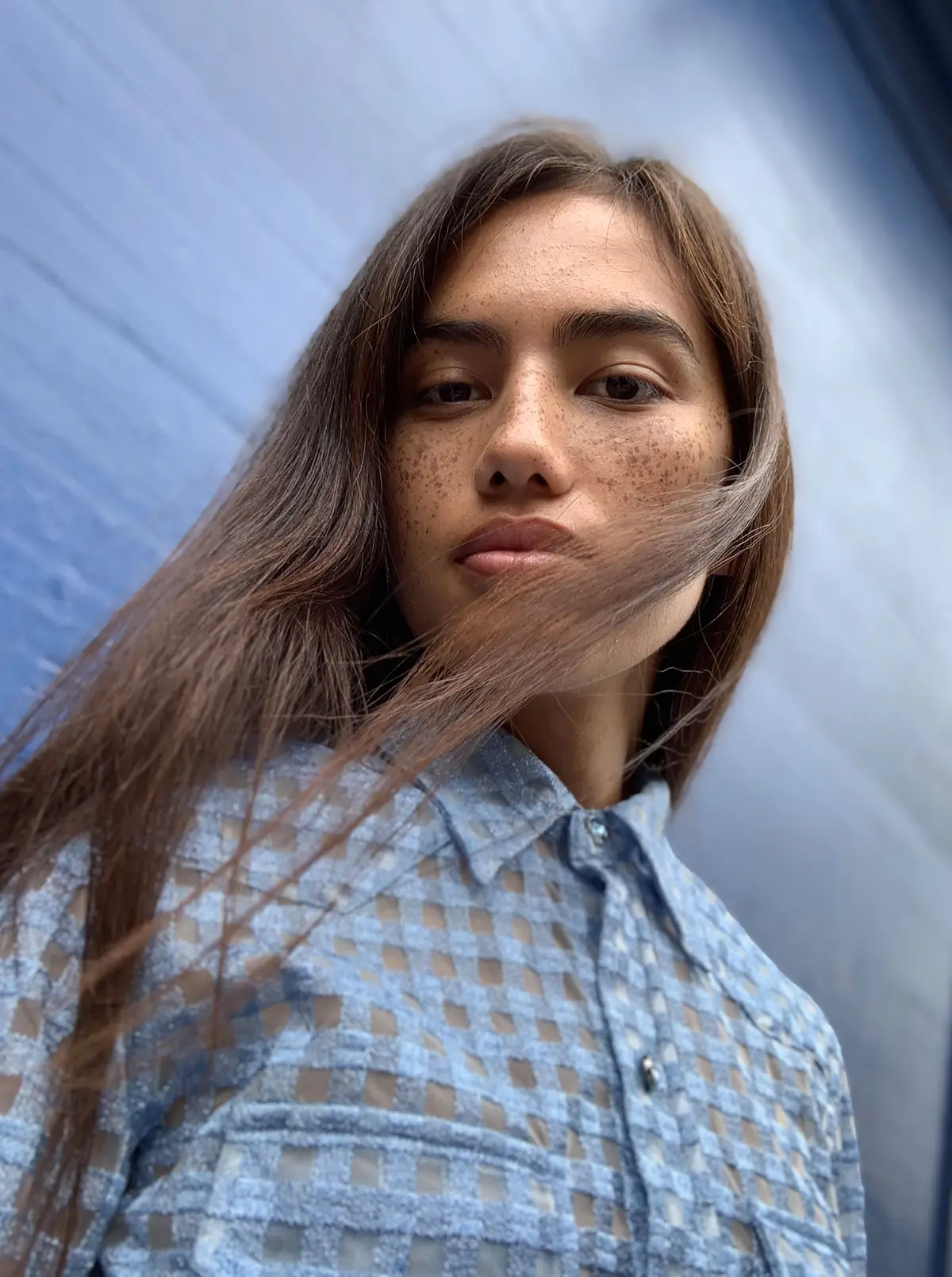 A young woman with freckles and long hair blowing across her face, against a blue wall
