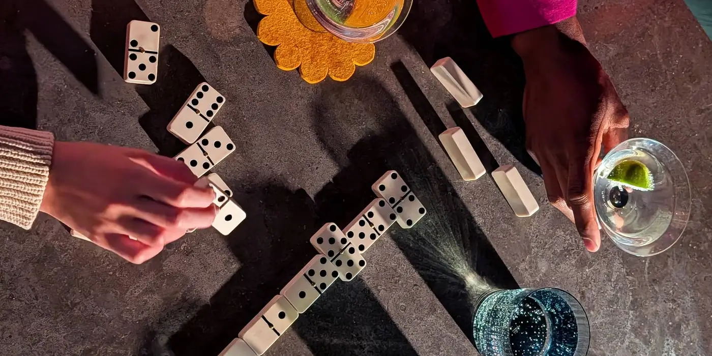 Wide: a top-down shot of hands playing dominoes beside drinks, the bubbles sharp