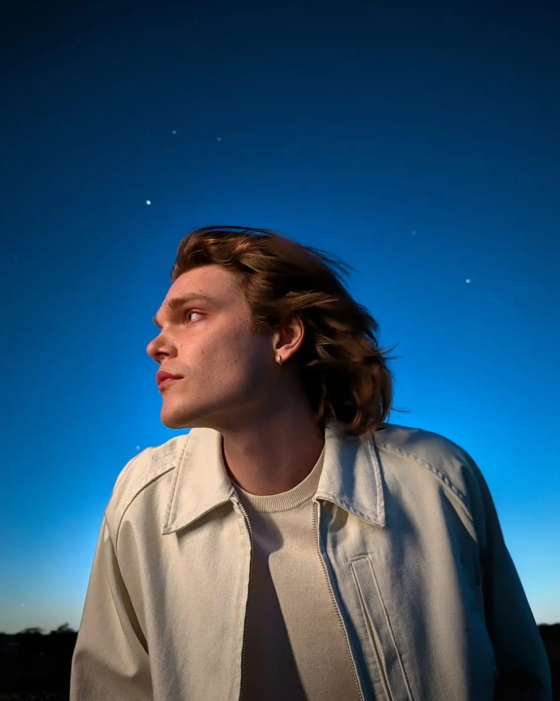 A man in a pale jacket looks up at a deep blue night sky with a few stars, his face clearly lit
