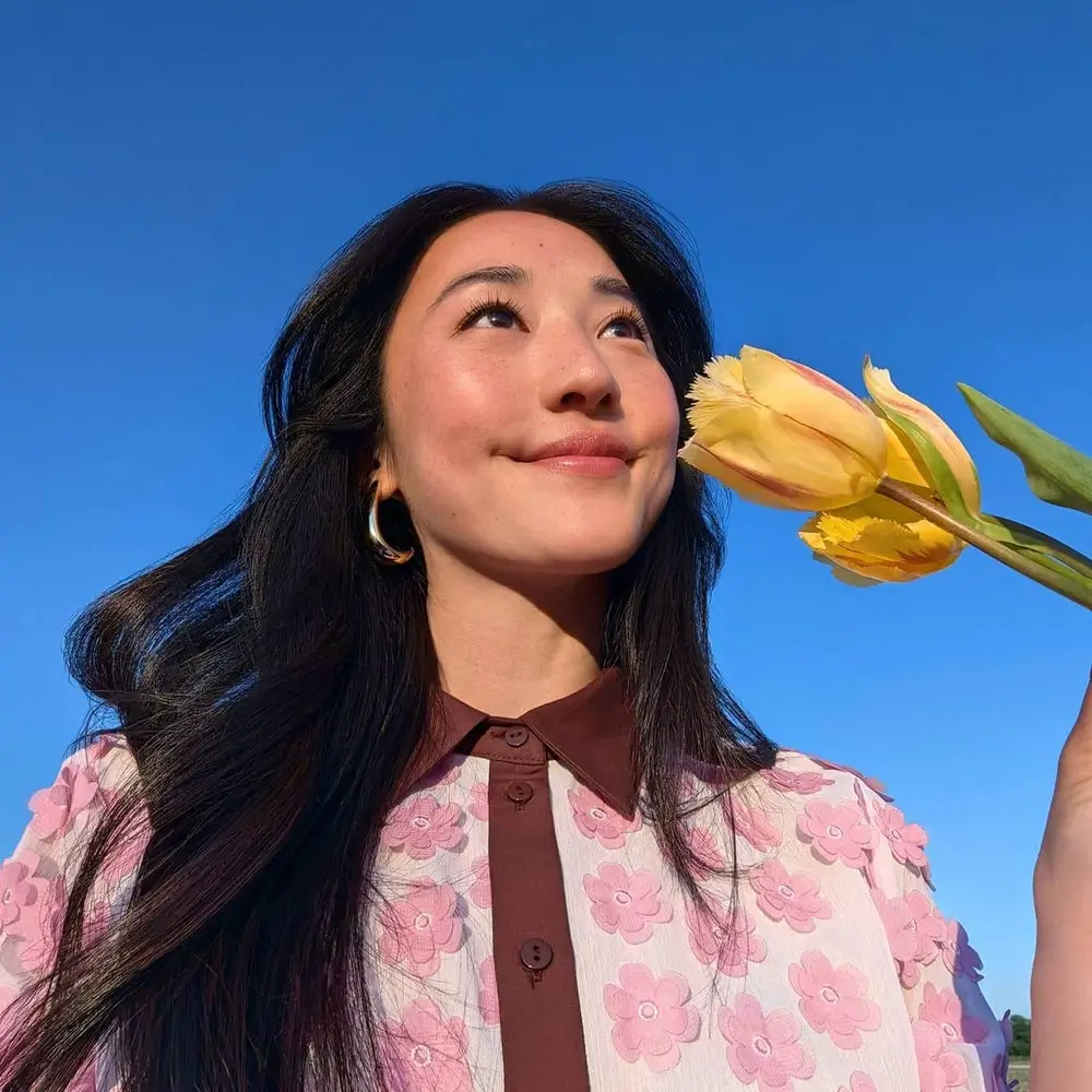 Front camera: a smiling woman holds a yellow tulip near her face under a blue sky