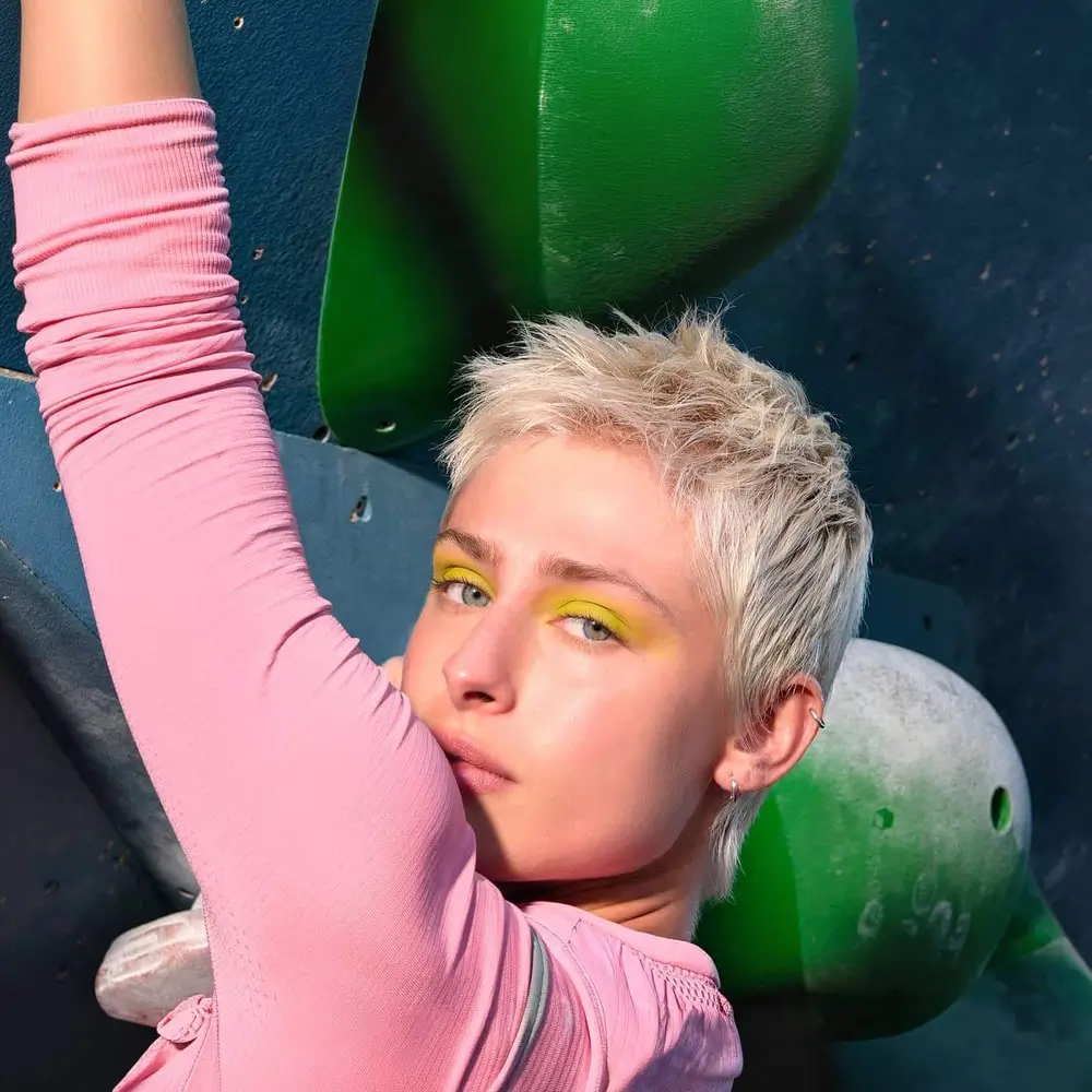 Telephoto: a woman with cropped silver hair and yellow eyeshadow, one pink sleeve raised against a climbing wall
