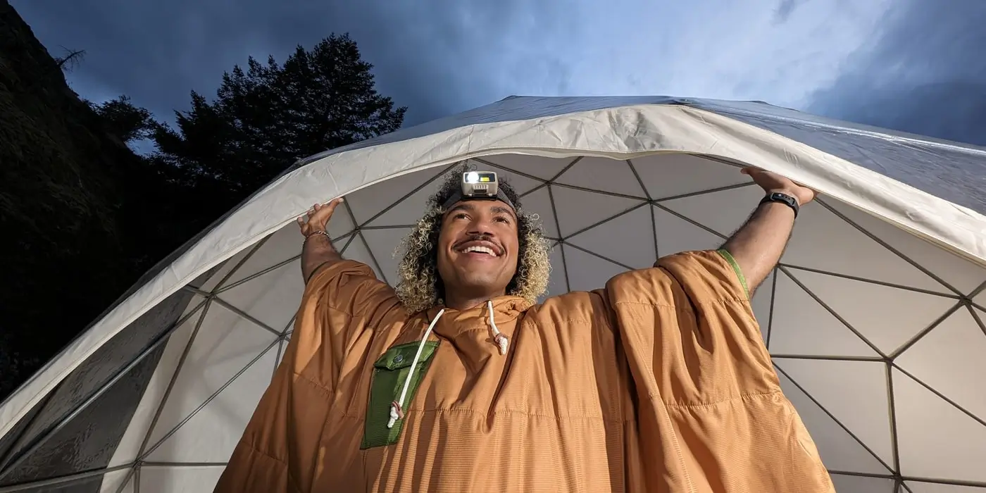 Main camera: a smiling man in an orange blanket stretches his arms under a dome tent at dusk