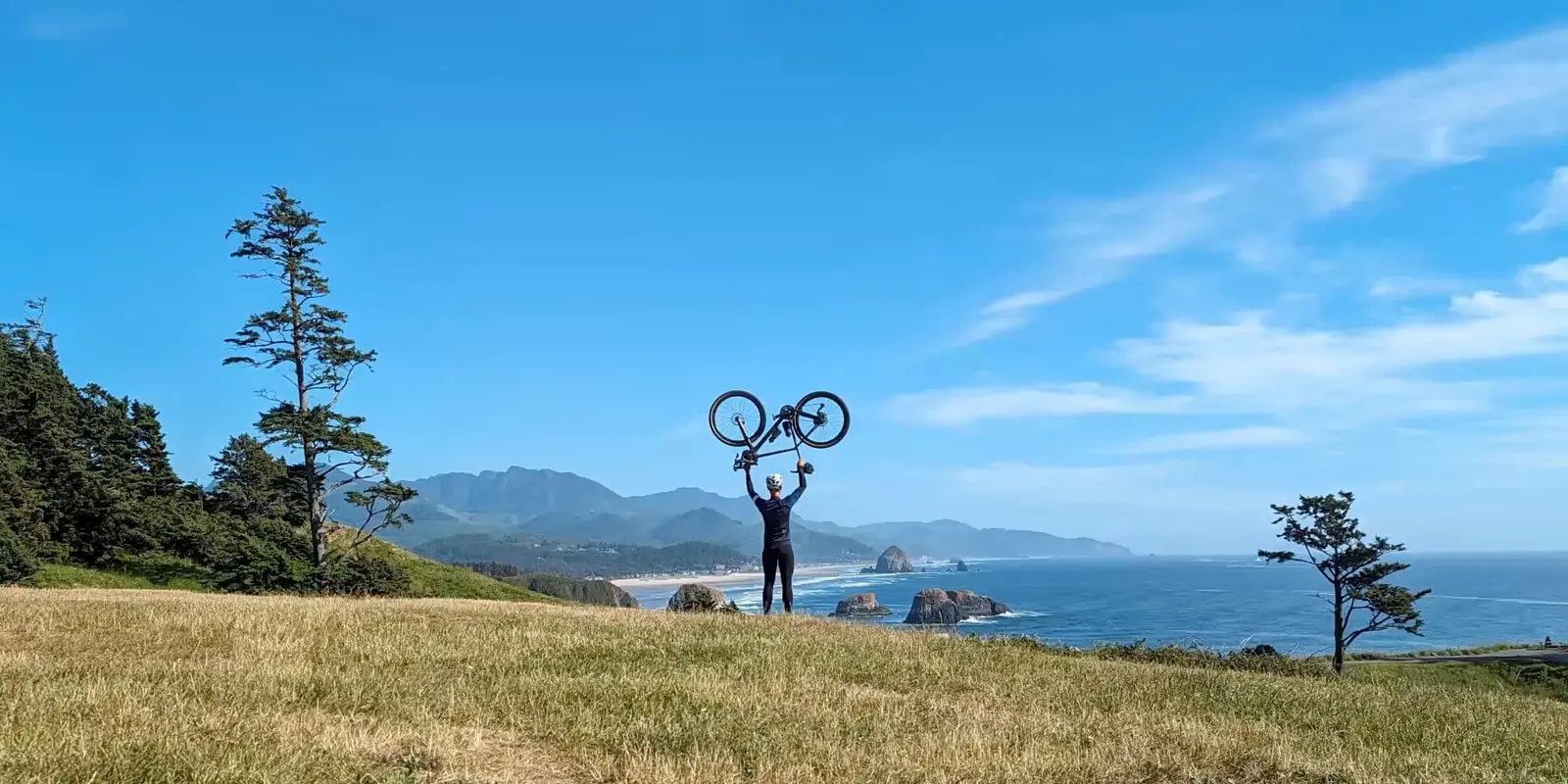 At 1x: a grassy clifftop above the sea and mountains, a cyclist small in the middle holding his bike overhead