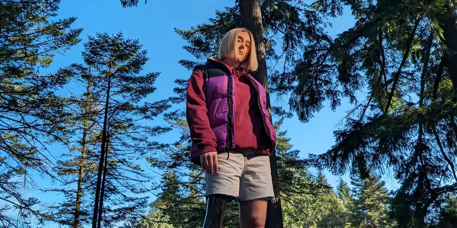At 1x: the woman in a purple and pink jacket from the knees up, pines against blue sky