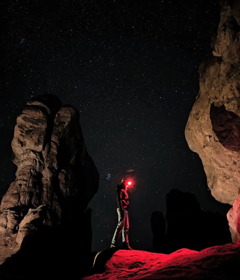 Night Sight: a person with a red head torch stands among rock towers under a starry sky