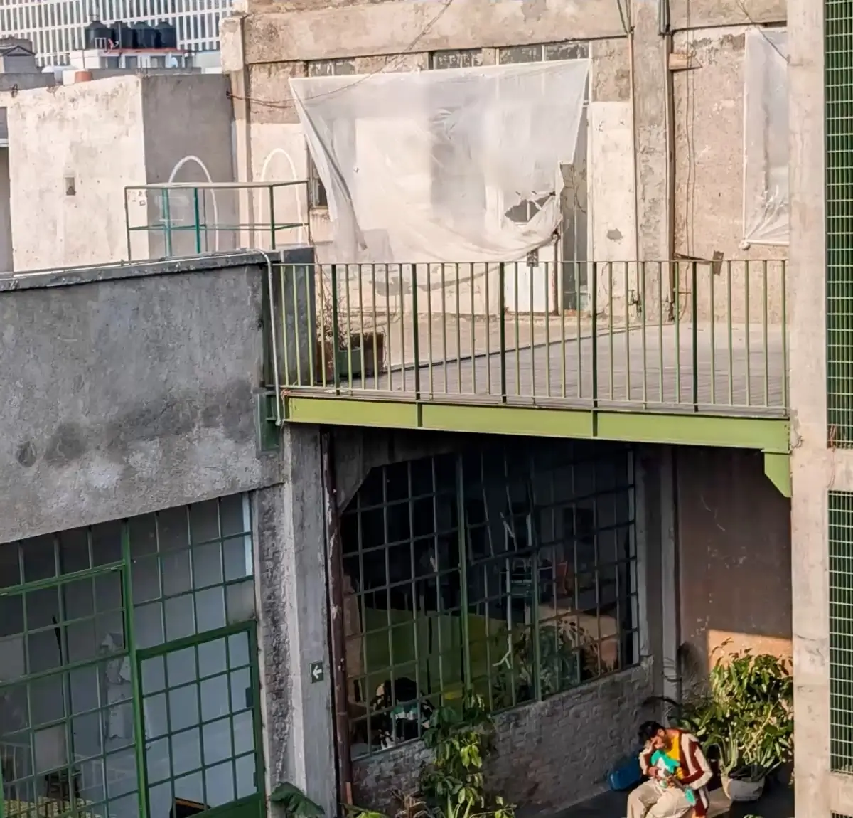 At 2x: the building's green-framed windows and balcony, the man sitting among plants