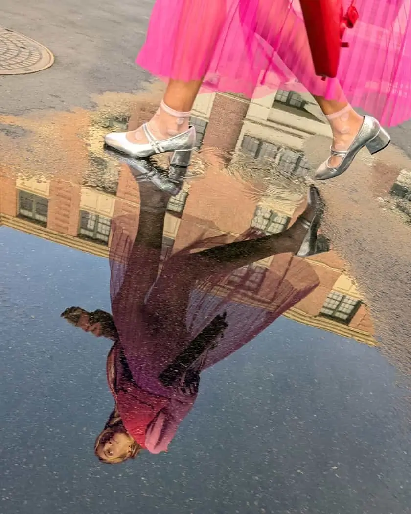 Main camera: a person in heels and a pink skirt steps through a puddle that reflects a building